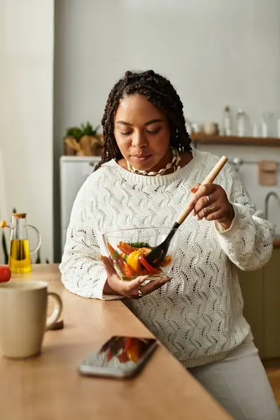A young African American woman prepares a vibrant salad in her cozy kitchen, savoring the moment. — Stock Photo