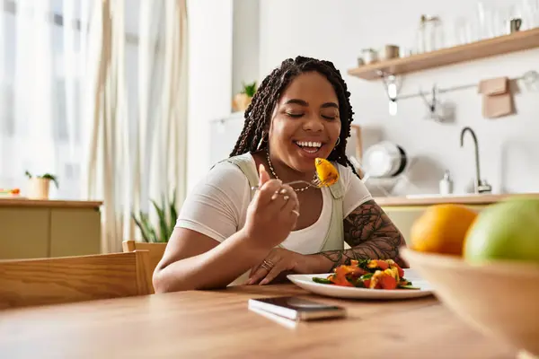 A young African American woman smiles joyfully while savoring her colorful salad at home. — Stock Photo