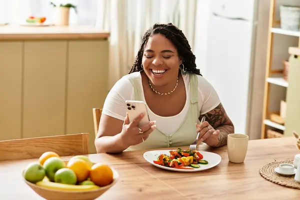 Uma jovem afro-americana sorri enquanto ela gosta de uma salada colorida e verifica seu telefone. — Fotografia de Stock
