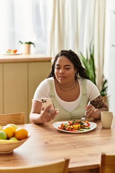 Uma jovem afro-americana senta-se em casa, saboreando uma salada vibrante e verificando seu telefone. — Fotografia de Stock