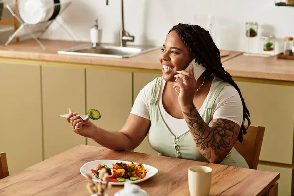 Uma jovem afro-americana sorri enquanto come sua salada fresca e fala ao telefone em casa. — Fotografia de Stock