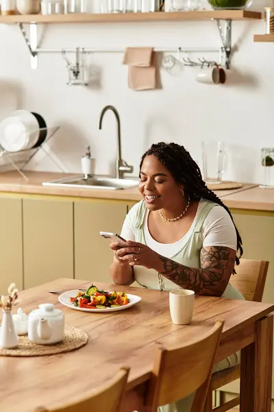 Uma jovem afro-americana senta-se em uma mesa de madeira, comendo uma salada colorida e mensagens de texto. — Fotografia de Stock