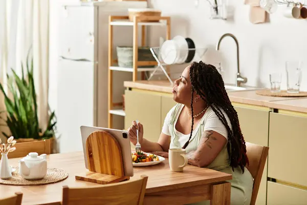 Uma jovem afro-americana saboreia uma salada fresca calmamente em sua acolhedora mesa de cozinha caseira. — Fotografia de Stock