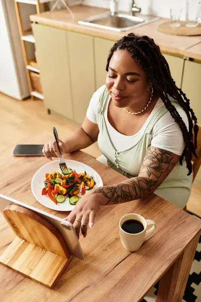 A young Black woman enjoys a colorful salad at her kitchen table while using her tablet. — Stock Photo