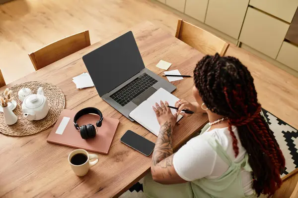 Una joven afroamericana trabaja a distancia en su mesa de comedor con herramientas para el éxito. - foto de stock