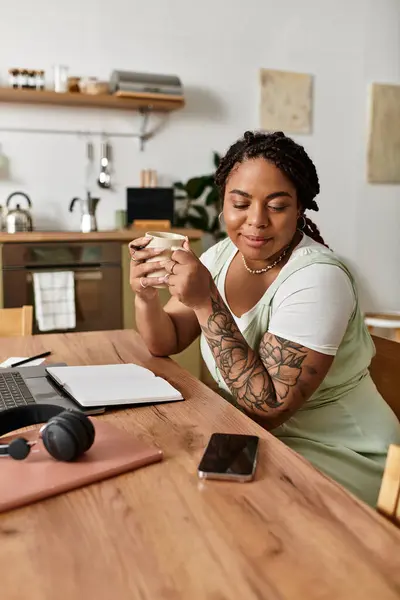 Mujer con el pelo trenzado sonríe mientras sostiene una taza, rodeada por su espacio de trabajo y su portátil. - foto de stock
