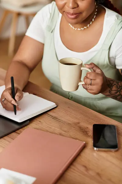 Une jeune femme concentrée sirote du café alors qu'elle écrit des notes dans son carnet à la maison. — Photo de stock
