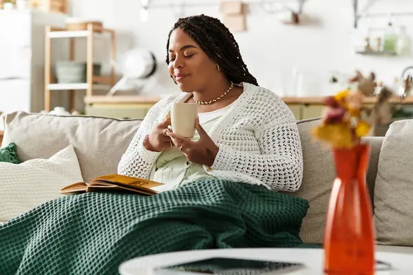 Una giovane donna afroamericana rilassata sorseggia tè su un divano mentre legge un libro nella sua casa. — Foto stock