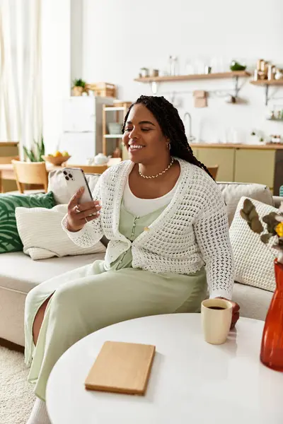 Eine junge Afroamerikanerin entspannt sich zu Hause, nippt am Kaffee und greift zum Handy. — Stockfoto