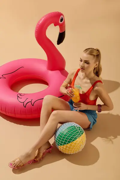 Beautiful young woman relaxes with a drink while seated beside a vibrant pool float. — Stock Photo