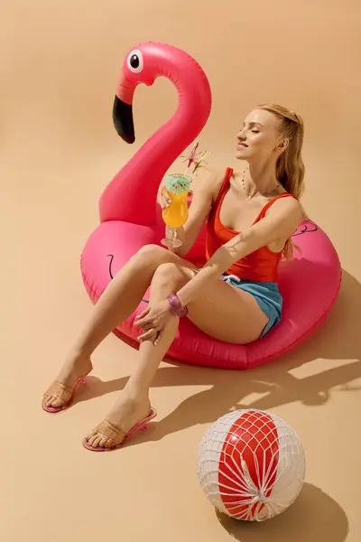 Entspannt auf einem lebhaften Flamingowagen nippt eine junge Frau in sommerlicher Glückseligkeit an einem farbenfrohen Getränk. — Stockfoto