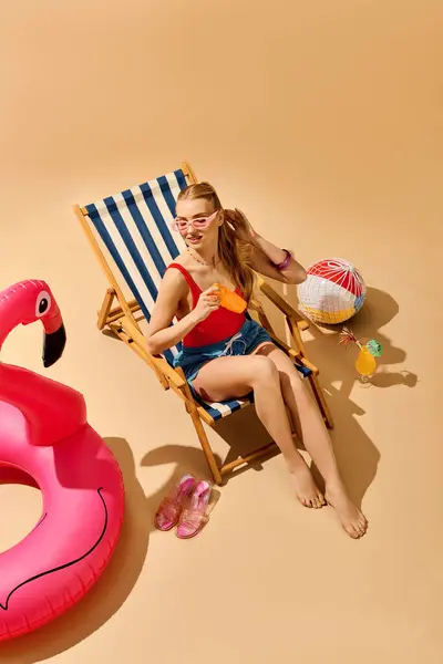 A young beautiful woman lounges on a stripy chair, sipping a drink under the warm sun at the beach. — Stock Photo