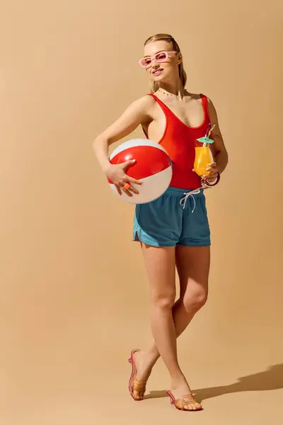 Young woman dressed in stylish swimwear holds a beach ball and a refreshing drink while smiling. — Stock Photo