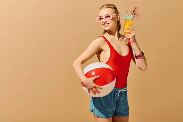 Una joven con un traje de baño rojo sonríe, sosteniendo una bebida y una pelota de playa, irradiando alegría. - foto de stock