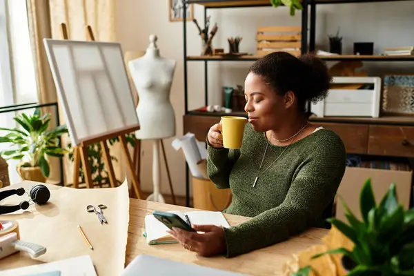 A beautiful woman enjoys a warm drink while surrounded by crafting materials in her workshop. — Stock Photo