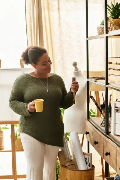 Una hermosa mujer disfruta de su oficio mientras sostiene un pincel y una bebida caliente en un taller. - foto de stock
