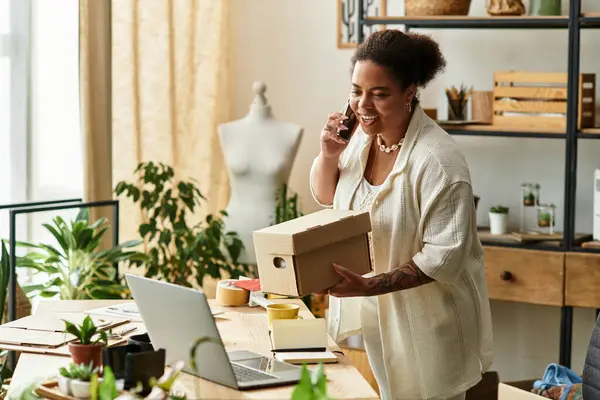 Una mujer sonriente habla por teléfono, sosteniendo una caja en un vibrante taller de elaboración. - foto de stock