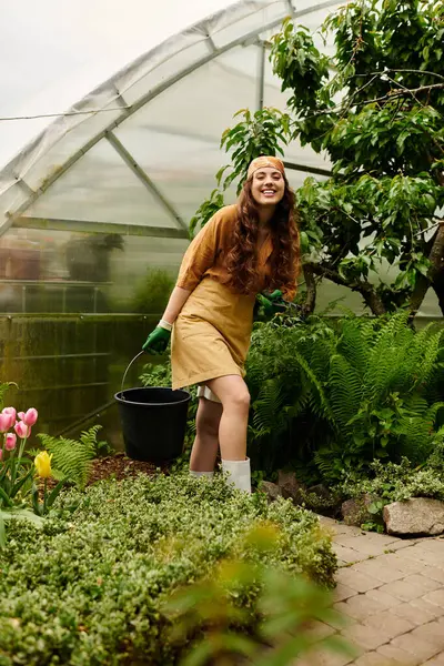 Um jardineiro alegre em um lenço de cabeça tende a plantas vibrantes em uma estufa exuberante, irradiando felicidade. — Fotografia de Stock