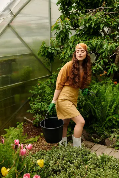 Uma jovem mulher em um lenço de cabeça nutre plantas vibrantes em uma estufa ensolarada, irradiando alegria. — Fotografia de Stock