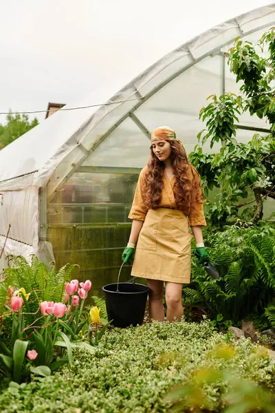 A joyful young woman in a headscarf tends to blooming flowers in her greenhouse garden. — Stock Photo