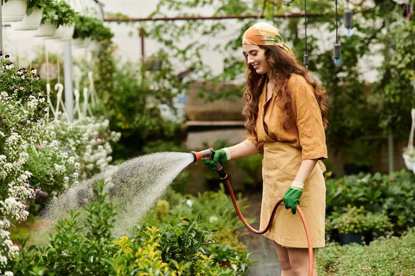 Uma jovem mulher em um lenço de cabeça feliz rega plantas em uma estufa vibrante. — Fotografia de Stock