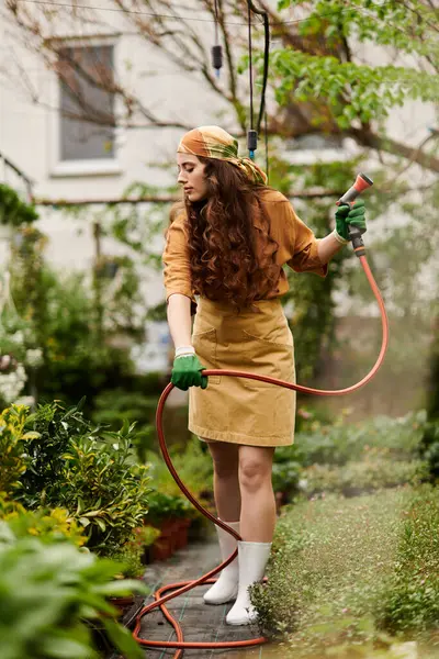 Um jardineiro vestindo um lenço de cabeça rega plantas em uma estufa exuberante, irradiando alegria e tranquilidade. — Fotografia de Stock