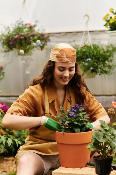 A happy woman in a headscarf works with soil and plants in a bright greenhouse, nurturing life. — Stock Photo