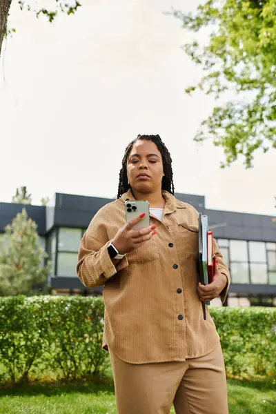 Educator interacts using a smartphone, holding books and strolling through a green campus area. — Stock Photo