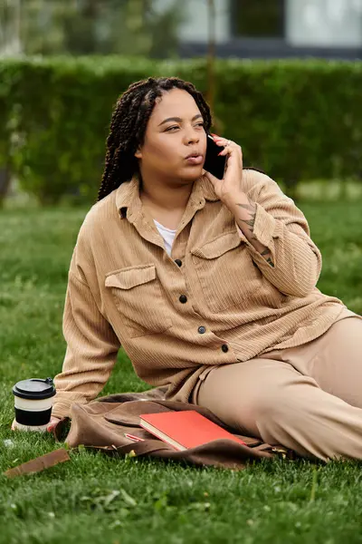 Mujer sentada en la hierba en el campus universitario, discutiendo por teléfono mientras sostiene una taza de café. - foto de stock