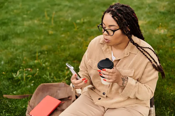 Una profesora universitaria se sienta en un campo cubierto de hierba, bebiendo café y mirando su teléfono mientras trabaja. - foto de stock