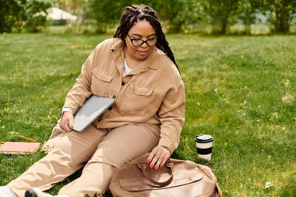 Une professeure d'université est assise sur l'herbe, à l'aide de son ordinateur portable, entourée de livres et de café. — Photo de stock
