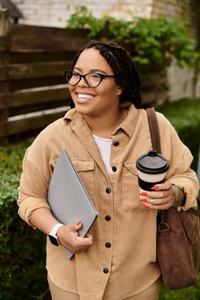 Une femme, professeur d'université, se promène à travers le campus, souriant et tenant une tasse de café et un ordinateur portable. — Photo de stock