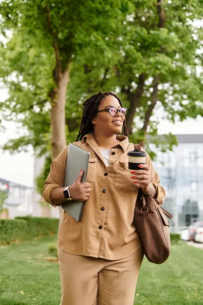 A university teacher strolls through campus, holding a laptop and coffee, smiling at the day. — Stock Photo