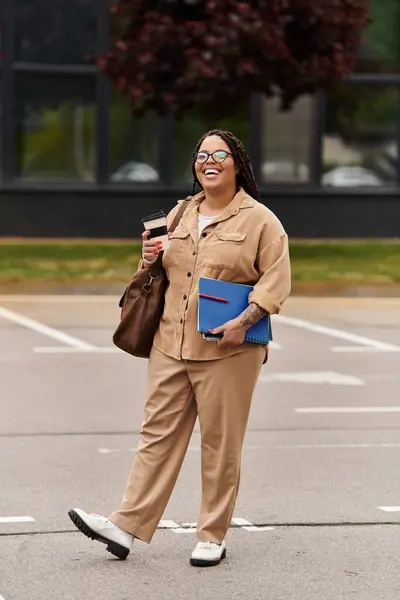 A cheerful university teacher carries coffee and books while walking outside on campus grounds. — Stock Photo