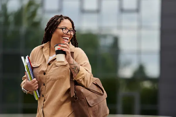 Une femme joyeuse porte des livres et un café, rayonnant d'enthousiasme sur un campus universitaire ensoleillé. — Photo de stock