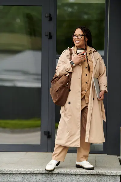 Confident woman in a trench coat stands outside a university, savoring a coffee and looking engaged. — Stock Photo