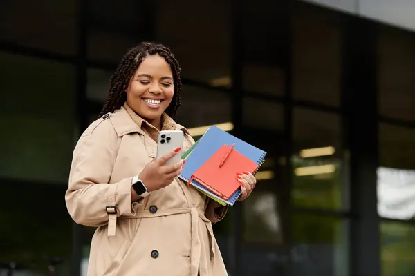 Donna fuori da un'università sorride al suo smartphone, tenendo quaderni colorati. — Foto stock