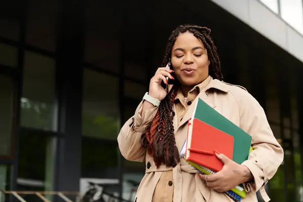 Una donna comunica al telefono mentre tiene dei quaderni fuori da un campus universitario. — Foto stock