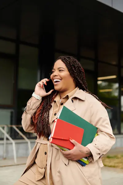 Woman with braids smiles while talking on the phone and holding colorful books on campus. — Stock Photo