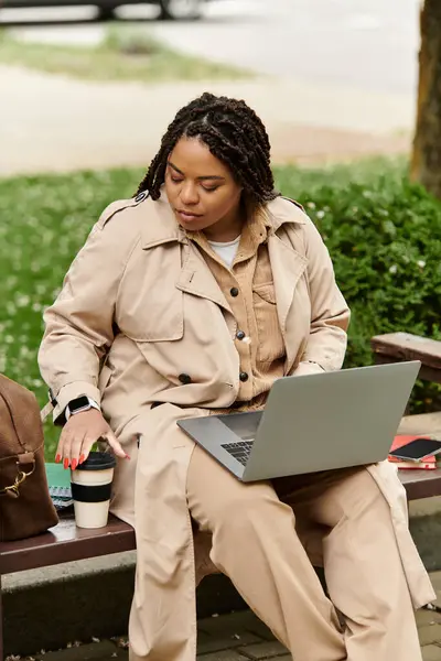 Donna in trench utilizza laptop e caffè mentre insegna all'aperto in un ambiente accogliente parco. — Foto stock