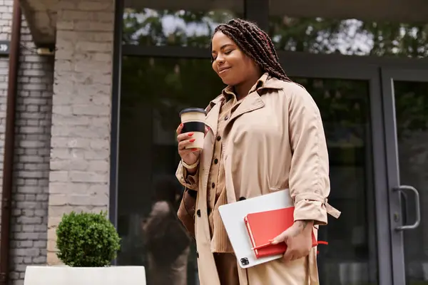 Femme dans une tenue élégante tient un café et un cahier, souriant dans un environnement universitaire. — Photo de stock