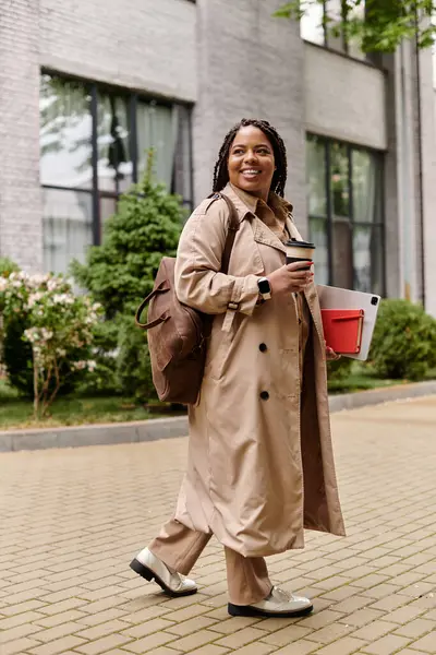 Una donna in un elegante trench passeggia per il parco universitario con libri e un caffè. — Foto stock