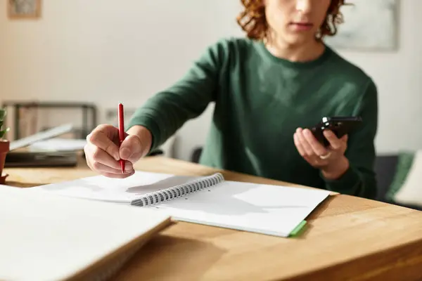 Um jovem senta-se em sua mesa, escrevendo notas com um lápis e usando seu telefone. — Fotografia de Stock