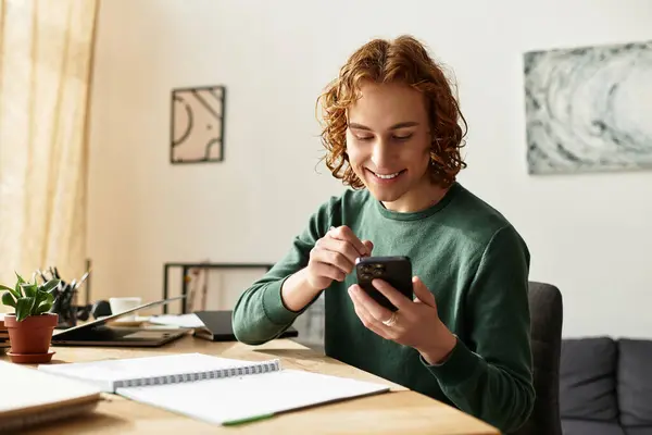 Un giovane uomo si gode il suo tempo a casa, usando felicemente il suo telefono in uno spazio di lavoro rilassato. — Foto stock
