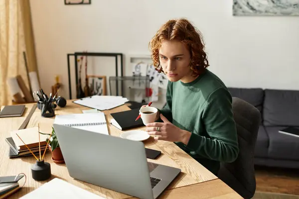 Un giovane uomo si concentra intensamente sul suo computer portatile sorseggiando caffè presso la sua accogliente scrivania di casa. — Foto stock