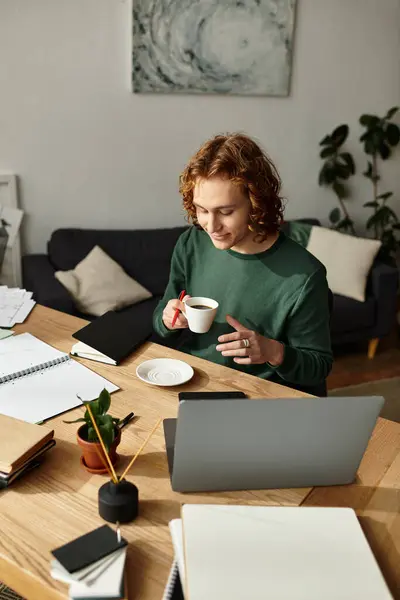 Young man smiles while sipping coffee, immersed in work at his cozy home workspace with plants. — Stock Photo