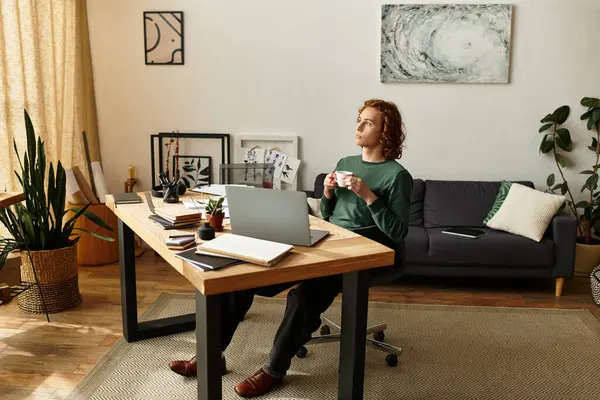 Young man sits at a modern desk, sipping a warm drink, surrounded by greenery and art at home. — Stock Photo
