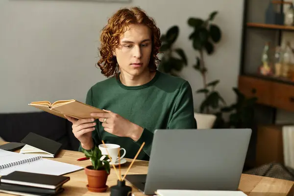 Un giovane uomo si siede comodamente nella sua accogliente casa, leggendo un libro mentre usa il suo computer portatile. — Foto stock