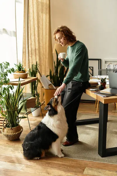 A young man enjoys coffee at home, playfully interacting with his dog among houseplants. — Stock Photo