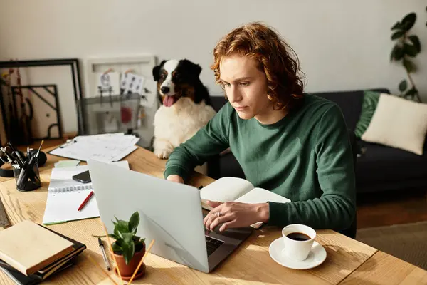 El joven se enfoca en su computadora portátil en casa mientras disfruta de una cálida taza de café y una compañía acogedora. - foto de stock
